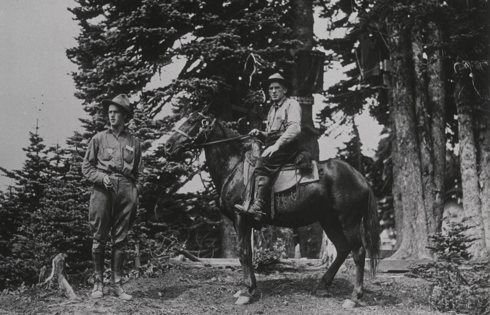 A man stands by another on horseback. Both wear breeches, boots, shirts, and flat brim hats. One has a round badge on his shirt.